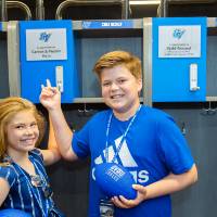 Two children holding footballs, pointing to a named locker at the Jamie Hosford Football Center dedication.